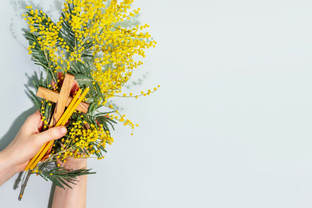 Easter religion card. Womans hands hold wooden crucifixion, mimosa sprig and church candles on blue background. Concept of Holy Week, Palm Sunday, Lent and religion holidays. Flat lay, copy spaceの写真素材