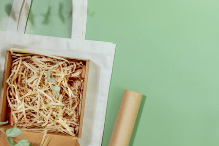 Eco friendly packaging concept. Cardboard box with shredded paper on textile shopping bag on green background with green leaves. Eco packaging goods from online stores and delivery. Copy spaceの写真素材