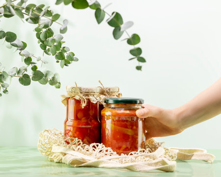 Probiotic foods. Sustainable still life with woman hand hold pickled or fermented vegetables. Lecho and ajvar in glass jars on mesh bag on blue background. Home food preserving or canningの写真素材