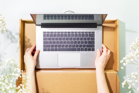 Buying and delivering new laptop. Romantic overhead shot of womans hands holding laptop in cardboard box on blue background with white spring flowers and shadows. Flat layの写真素材