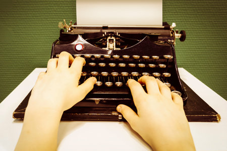 Hands of anonymous young person are typing on vintage typewriter filled with white blank sheet of...