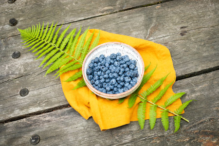 Bowl Of Fresh Blueberries On Yellow Cloth With Fern Leaves Over Rustic Wooden Table. Concept Of Forest Harvest, Healthy Food, Eco Products And Seasonal Aesthetics For Eco Visual Contentの写真素材