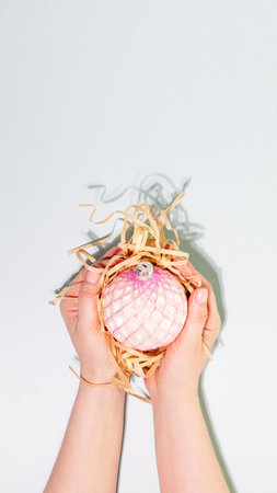 Female Hands Holding Pink Christmas Ornament In Eco-Friendly Packaging With Straw Filler. Concept Of...