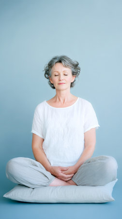 Senior Woman Sitting Cross Legged On Cushion In Minimal Blue Studio Practicing Mindfulness Meditation. Concept Of Healthy Aging, Stress Relief, And Calm Lifestyle Inspirationの素材