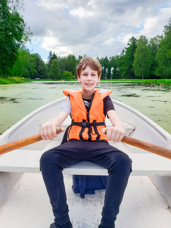Summer vacations. Boy in life jacket rows boat on tranquil lake, capturing essence of family vacations focused on outdoor activities and promoting healthy lifestyle among youth. Vertical formatの写真素材