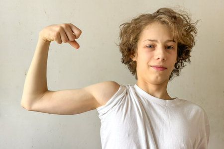 Confident Teen Boy Flexing Arm Indoors During Daytime For Concepts Of Body Positivity, Self-Esteem, Youth Identity, Physical Growth, And Empowering Visuals For Mental And Emotional Strength Campaignsの写真素材