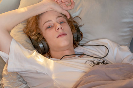 Teen Boy Lying In Bed With Headphones In Warm Evening Light. Concept of Teen Mental Health Support,...