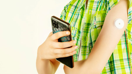 Teen holding smartphone with glucose sensor on arm against light background