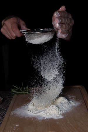 Woman chef hands holding a sieve with flour falling on wooden board with rosemary on the table.の写真素材