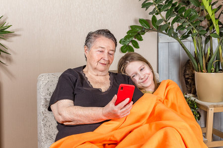Happy grandmother and cute granddaughter use mobile phone together while sitting on the sofa, smiling elderly grandmother and little girl have fun taking selfie on the phone, copy space, happiness conceptの写真素材