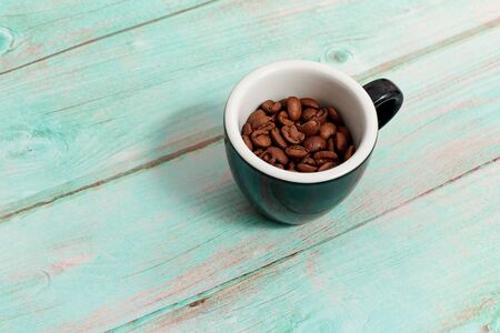a small Cup with brown roasted coffee beans on a light blue wooden background close-up. the Cup is half empty, white inside and black outside side view. some coffee beans in a Cupの写真素材