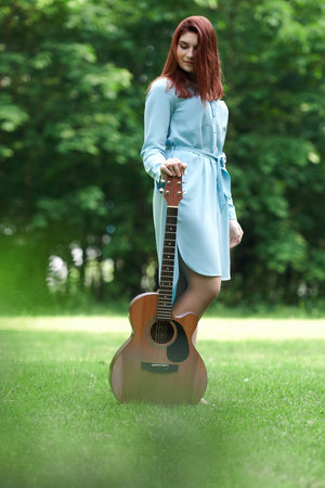 a girl with a guitar stands in an open green area. a full-length girl with a guitar is on the lawn...