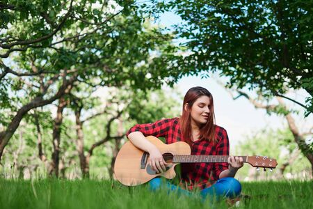 a girl in a checked shirt and jeans is sitting on a green lawn, playing a classical acoustic guitar with her own hands and singing. A girl in nature plays a musical insの写真素材