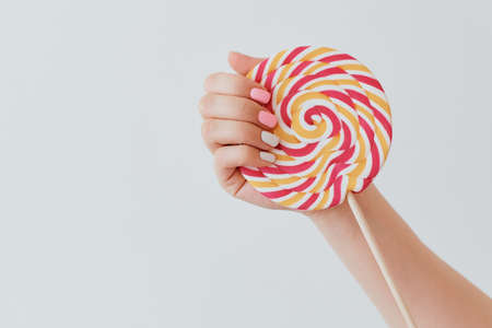 a hand with a pink and white manicure holds a caramel round candy on a stickの写真素材