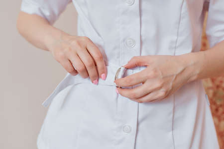 women's hands tighten the belt on a white medical coat with buttons. a woman puts on a cosmetologist's robeの写真素材