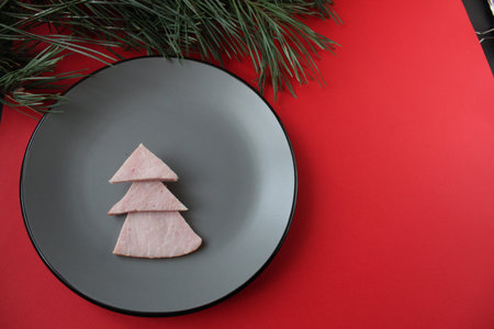 edible Christmas tree and sausages on a gray plate on a red background with a branch of a Christmas...