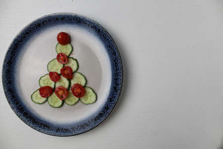 edible Christmas tree of cucumber vegetables and tomato on a white plate with a blue rim on a white gray background with space for text.の写真素材
