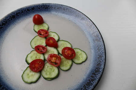 edible Christmas tree of cucumber vegetables and tomato on a white plate with a blue rim on a white gray background with space for text.の写真素材