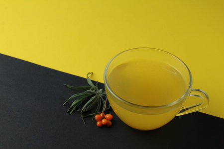 sea buckthorn tea drink on a black yellow bright background. Sea buckthorn drink tea juice in a glass cup next to a spout of sea buckthorn leaves orange berries on a black and yellow background. Creative creative photography. Seasonal berry.の写真素材