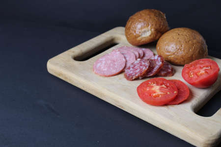 preparation for cooking. Ingredients for salad sandwich. Vegetables tomatoes bread bun buckwheat sausage ham lie on a tray board on a black background with copyspace for text.の写真素材