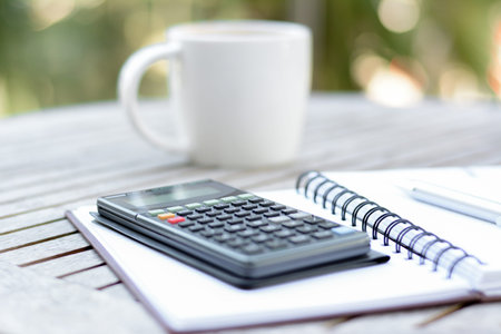 Calculator & pen over copybook on wood table with blurred coffee cup background - soft focus style