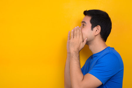 Young man shouting with cupped hands around mouth, yellow background studio shot with cocy space