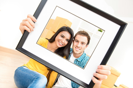 Happy interracial couple smiling through picture frame after moving into new houseの写真素材