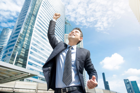 Businessman smiling and raising his fist in the air, with office building background - business success, achievement, and win conceptsの写真素材