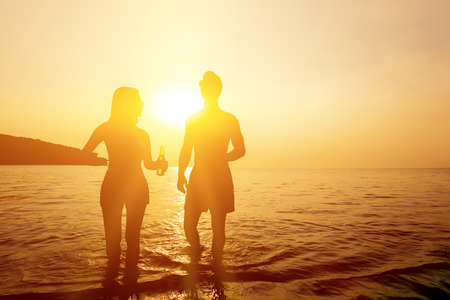 Silhouette of couple holding beer bottles walking in seawater at the beach in twilight sunsetの写真素材