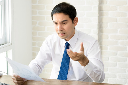 Asian businessman getting upset and disappointed after reading document at his working deskの写真素材