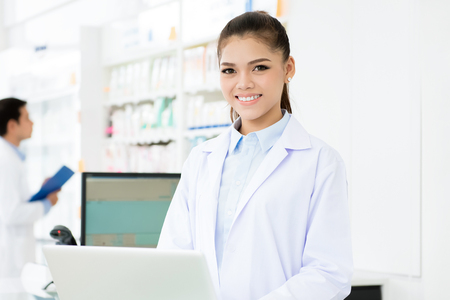 Young smiling Asian woman pharmacist in white gown coat uniform working on laptop computer in chemist shop or pharmacyの写真素材