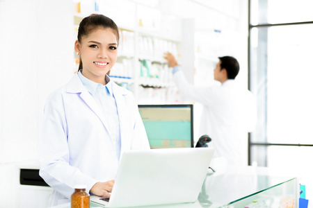 Young smiling Asian woman pharmacist in white gown coat uniform working on laptop computer in chemist shop or pharmacyの写真素材