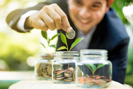 Businessman putting coin into the glass jar with young plant, demonstrating financial growth through saving plans and investment schemesの写真素材