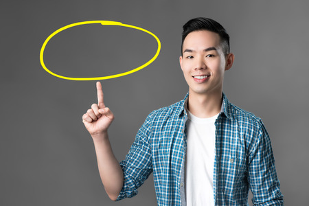 Cheerful smiling young Asian man pointing his finger up to empty circle - can put your texts insideの写真素材