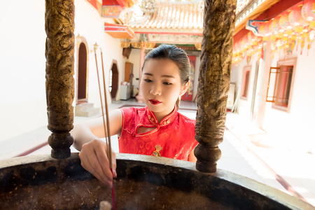 Asian woman in red qipao dress praying with incense sticks during Chinese or Lunar new year in the templeの写真素材