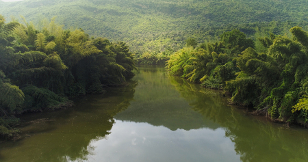 Beautiful natural scenery of river in tropical green forest with trees on two sides and mountain in backgroundの写真素材