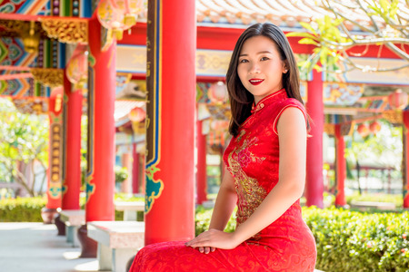 Smiling Asian woman in traditional red cheongsam qipao dress sitting with hands resting on lap in beautiful Chinese templeの写真素材