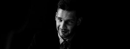 Sly man in formal business suit staring at interlocutor in dark shadow banner background, black and...