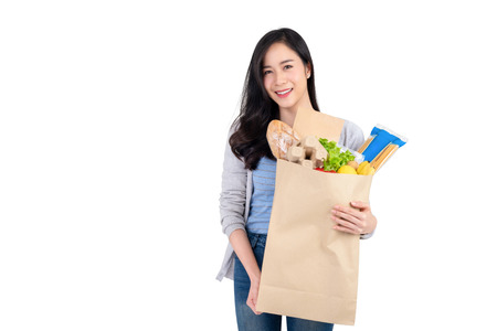 Beautiful smiling Asian woman holding paper shopping bag full of vegetables and groceries, studio shot isolated on white background