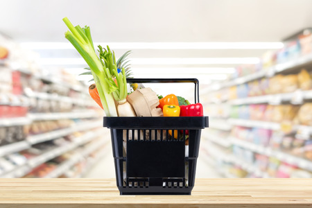 Food and groceries in black shopping basket on wood table with blurred suppermarket aisle in backgroundの写真素材
