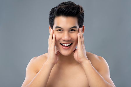 Portrait of handsome happy smiling young Asian man with hands touching face studio shot isolated on gray backgroundの写真素材