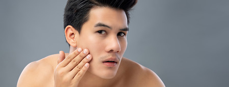 Portrait of shirtless young handsome Asian man checking his face for skin care and beauty concepts, studio shot isolated on gray banner background with copy spaceの写真素材