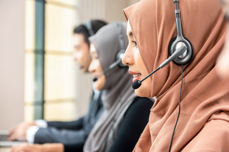 Muslim woman wearing microphone headset working as customer care operator with team in call center...