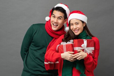 Happy smiling young Asian couple wearing colorful red and green sweaters with Christmas gift boxes studio shot on gray backgroundの写真素材