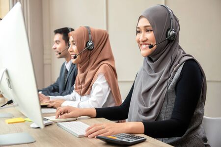 Smiling Asian muslim woman wearing microphone headsets working as customer care operator with team in call center officeの写真素材