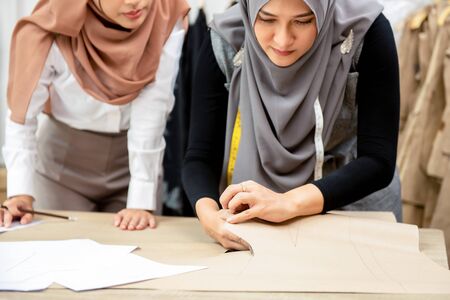 Muslim woman fashion designer working together with her colleague cutting clothing pattern in tailor shopの写真素材
