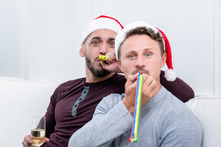 Playful gay couple blowing party horns while sitting on the couch celebrating Christmas together at homeの写真素材