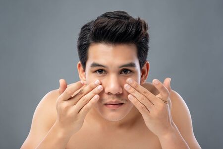 Portrait of shirtless young handsome Asian man checking his face for skin care and beauty concepts, studio shot isolated on gray backgroundの写真素材