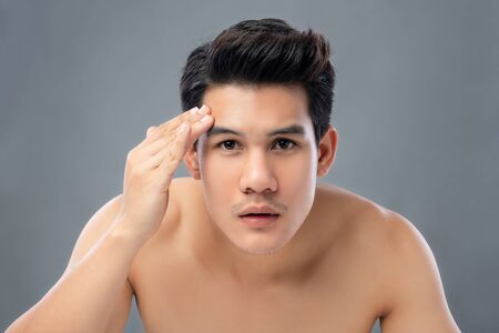 Portrait of shirtless young handsome Asian man checking his face for skin care and beauty concepts, studio shot isolated on gray backgroundの写真素材