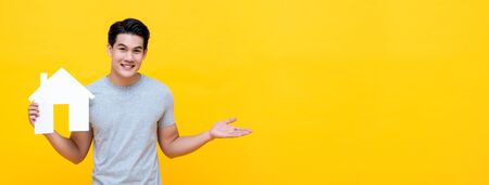 Portrait of young handsome Asian man holding paper home on yellow banner background for real estate conceptの写真素材